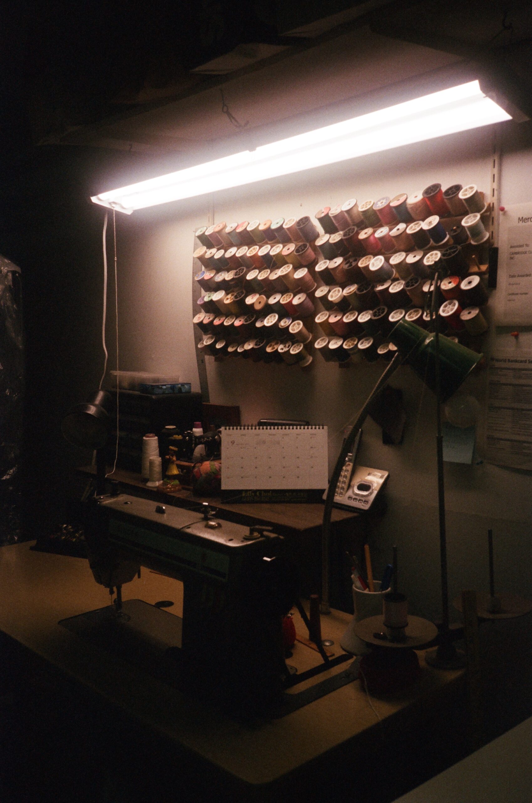 A sewing machine sits beneath a wall of thread and a florescent light