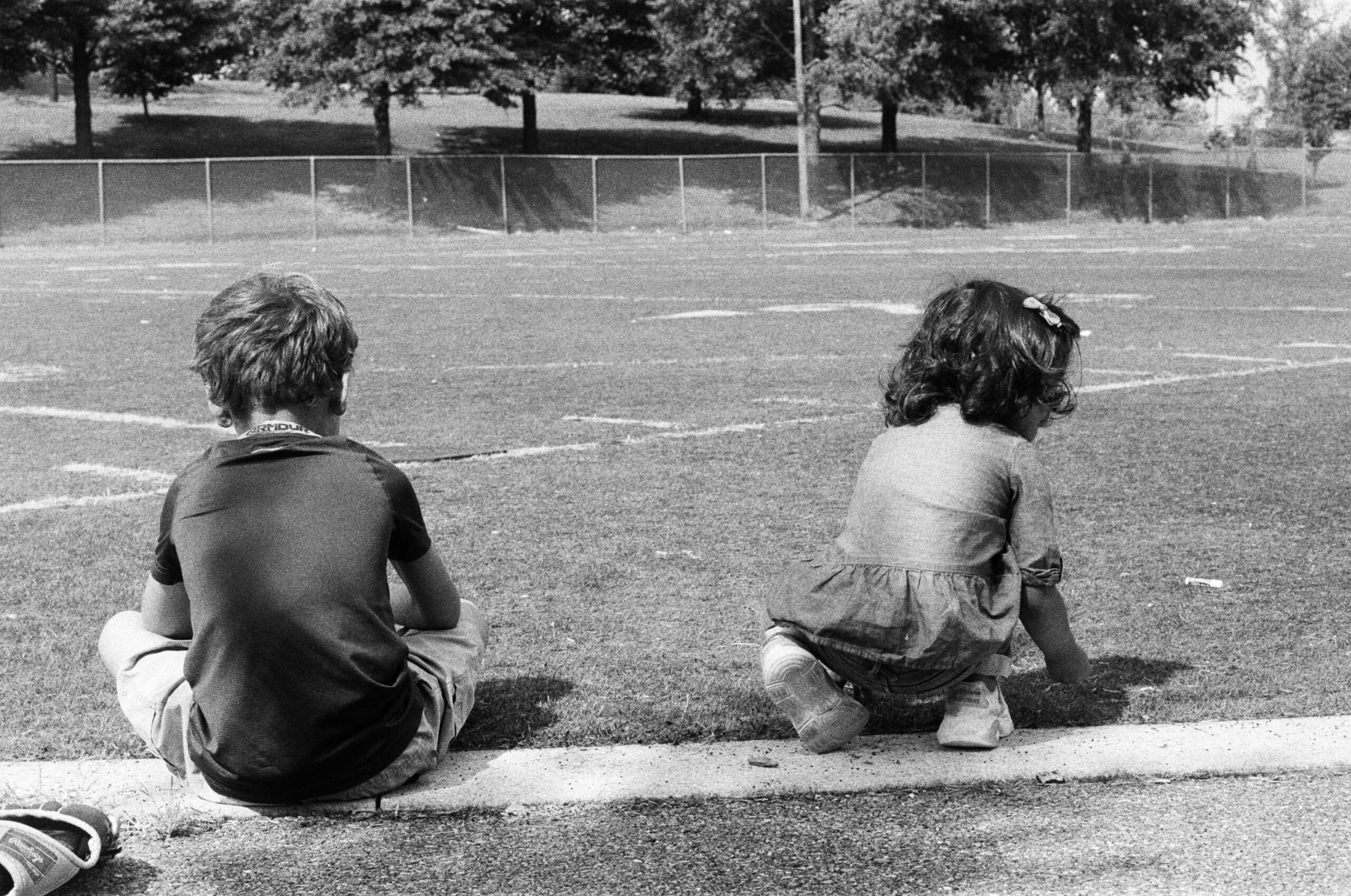 Two children sit in a field with their backs to the camera, picking grass