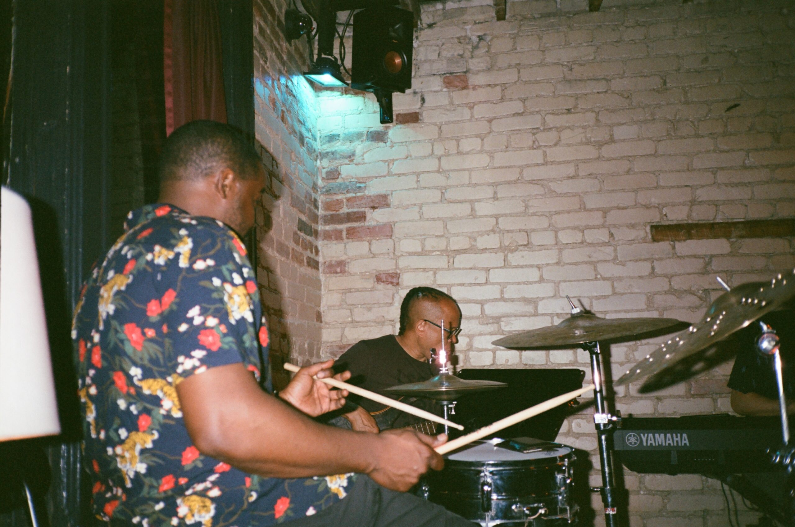 a drummer and bassist play in front of a white-washed brick wall